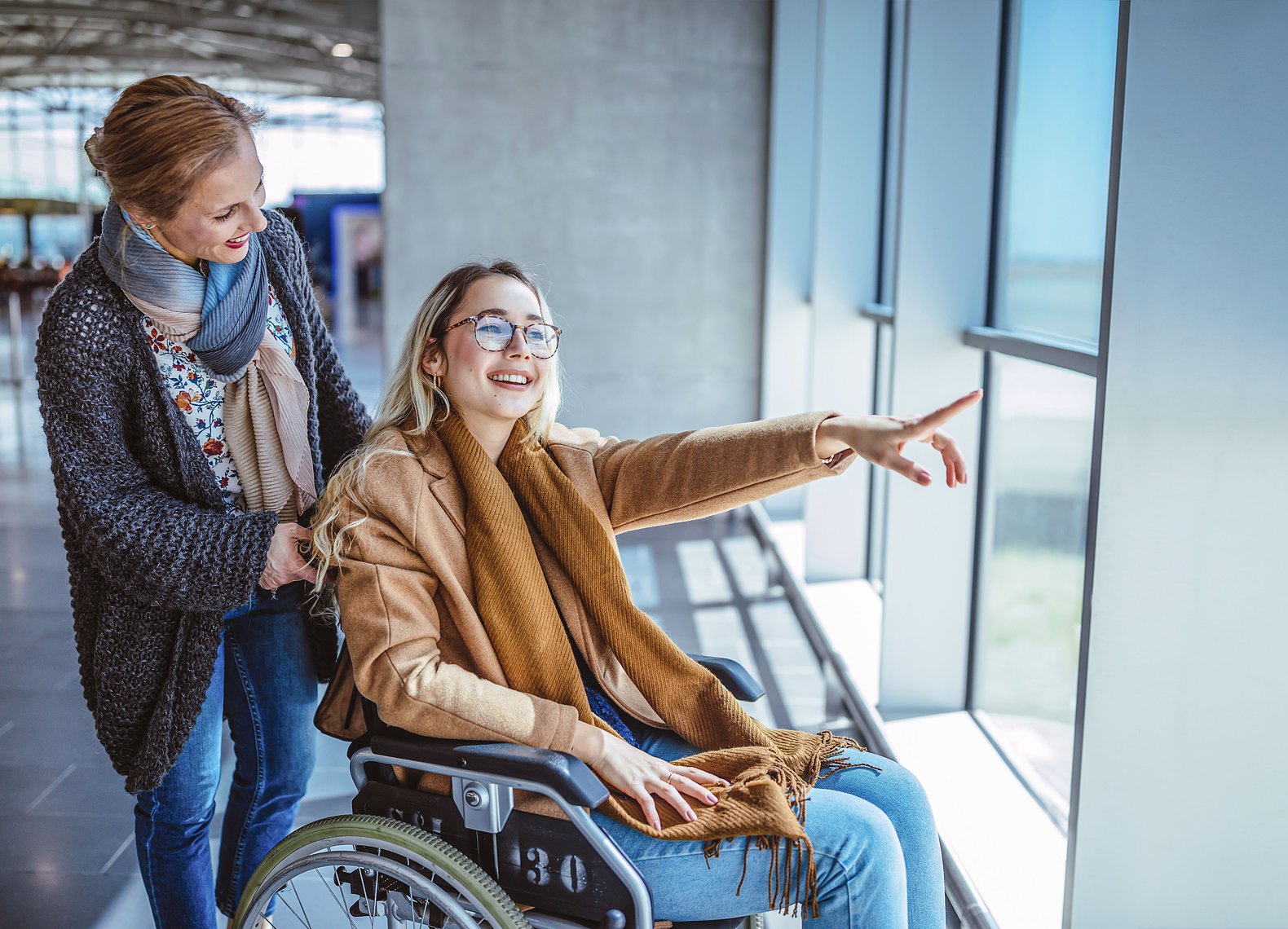 Eine Rollstuhlfahrerin schaut zusammen mit ihrer Begleiterin fröhlich aus dem Fenster einer Bahnhofshalle.