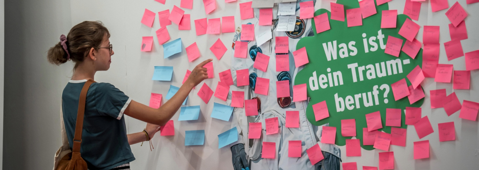 Eine junge Frau klebt ein Post-It an eine Wand, auf der bereits viele Post-Its hängen.