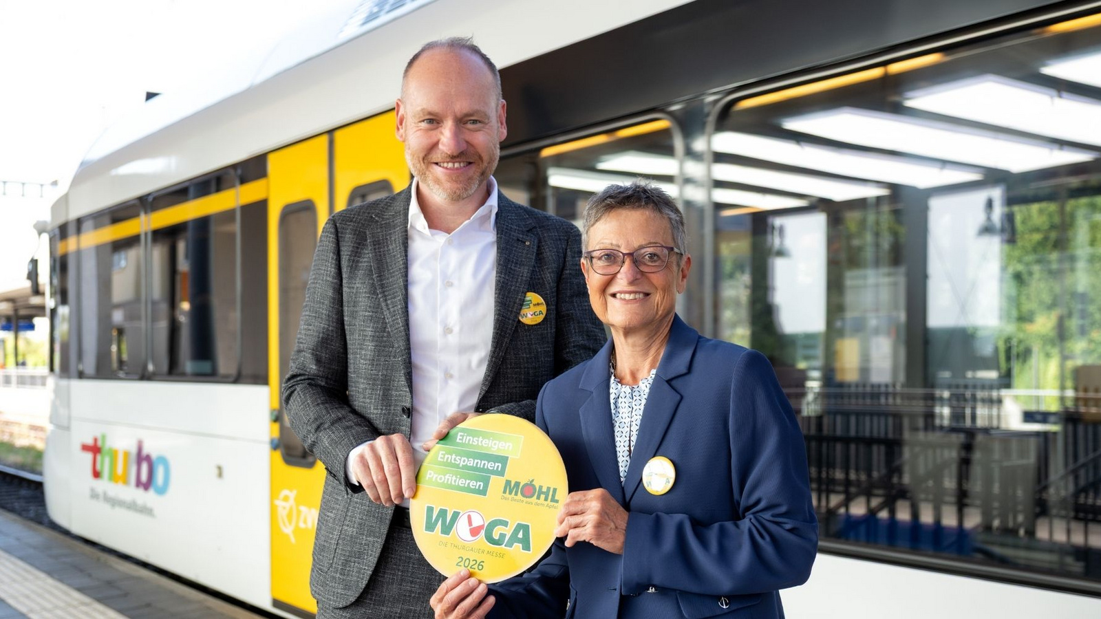 WEGA-Präsident Beat Bollinger und Thurbo Unternehmensleiterin Claudia Bossert halten den gelben WEGA-öV-Button vor einem Thurbo Zug in die Kamera.