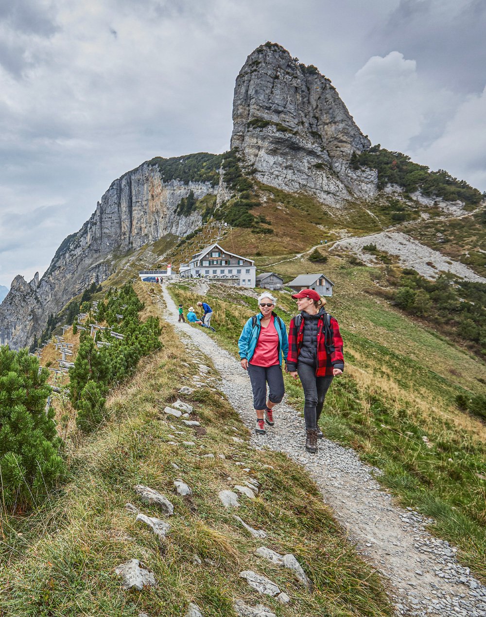 Zwei Wanderinnen laufen auf einem schmalen Berg-Pfad dem Betrachter bzw. der Betrachterin entgegen.