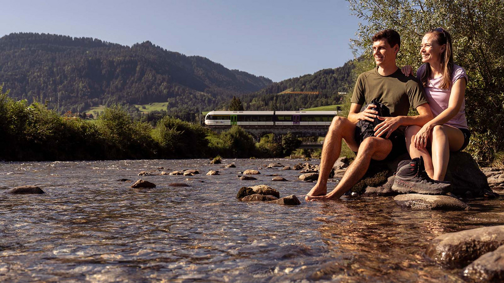 Paar sitzt an einem Fluss, im Hintergrund fährt ein Thurbo Zug über Brücke.