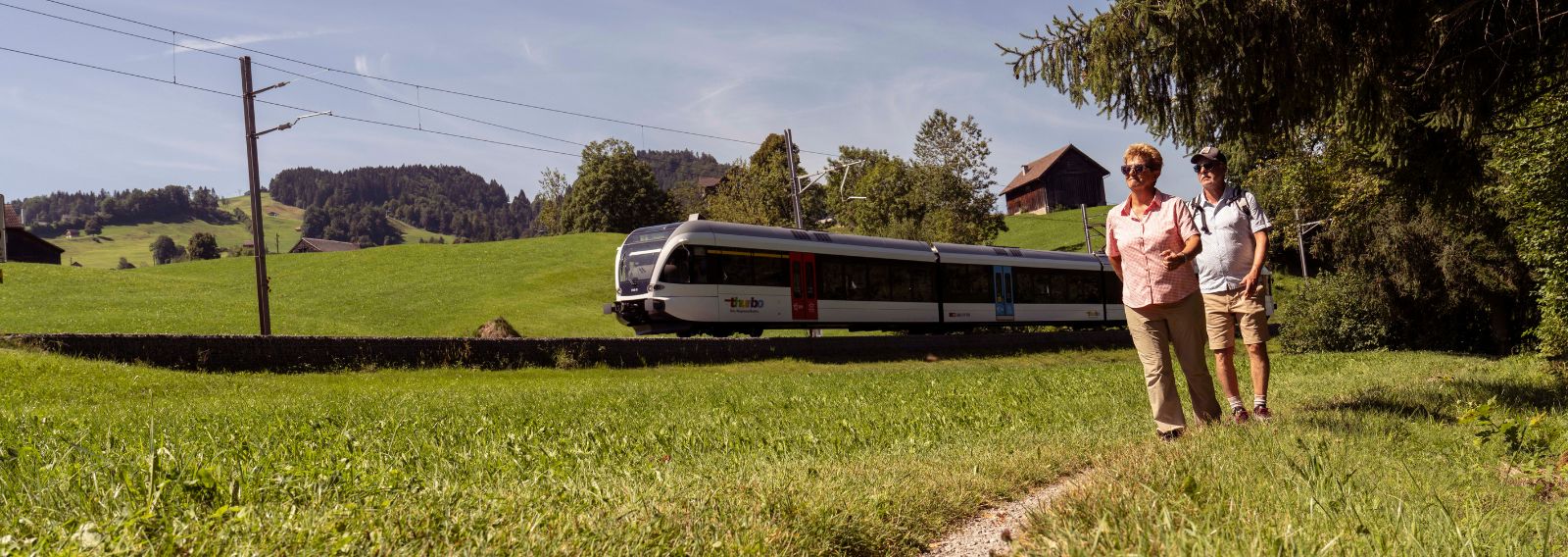 Zwei Menschen wandern durch die grüne Landschaft, ein Thurbo Zug fährt im Hintergrund.